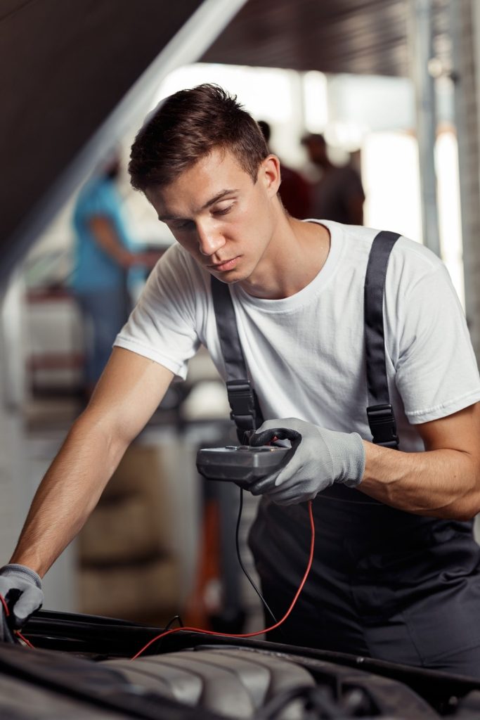 A young man in a white shirt and gray overalls is working on a car engine, using a device with wires in his hand.